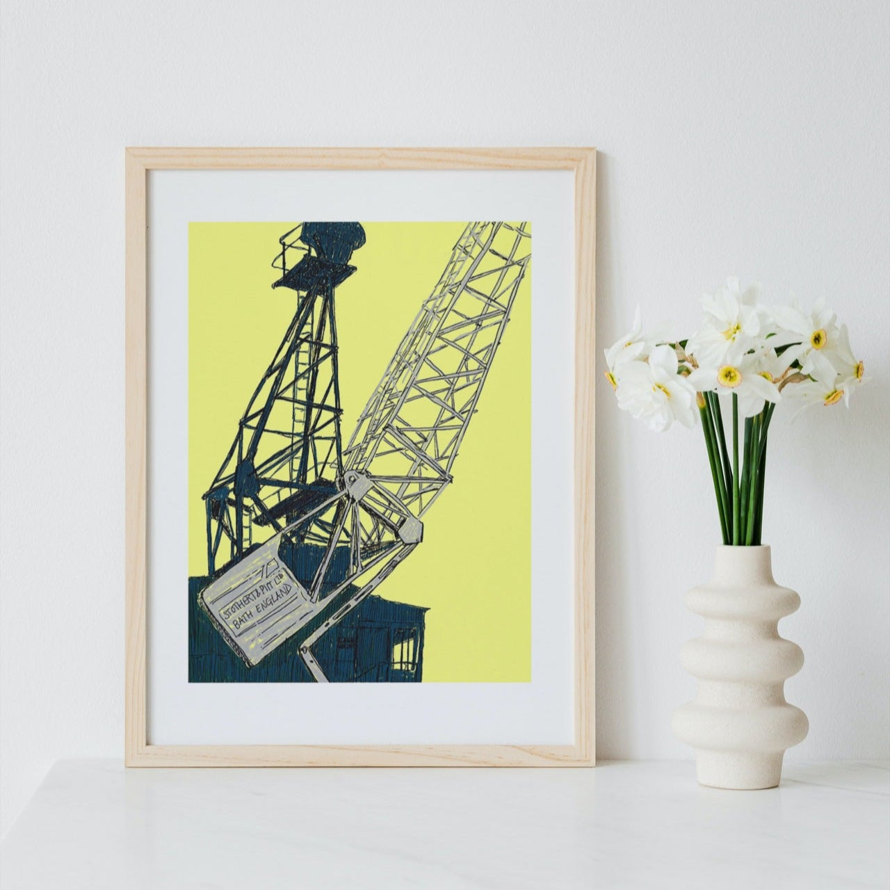 Framed artwork of a crane on a yellow background next to a vase with flowers on a white surface.