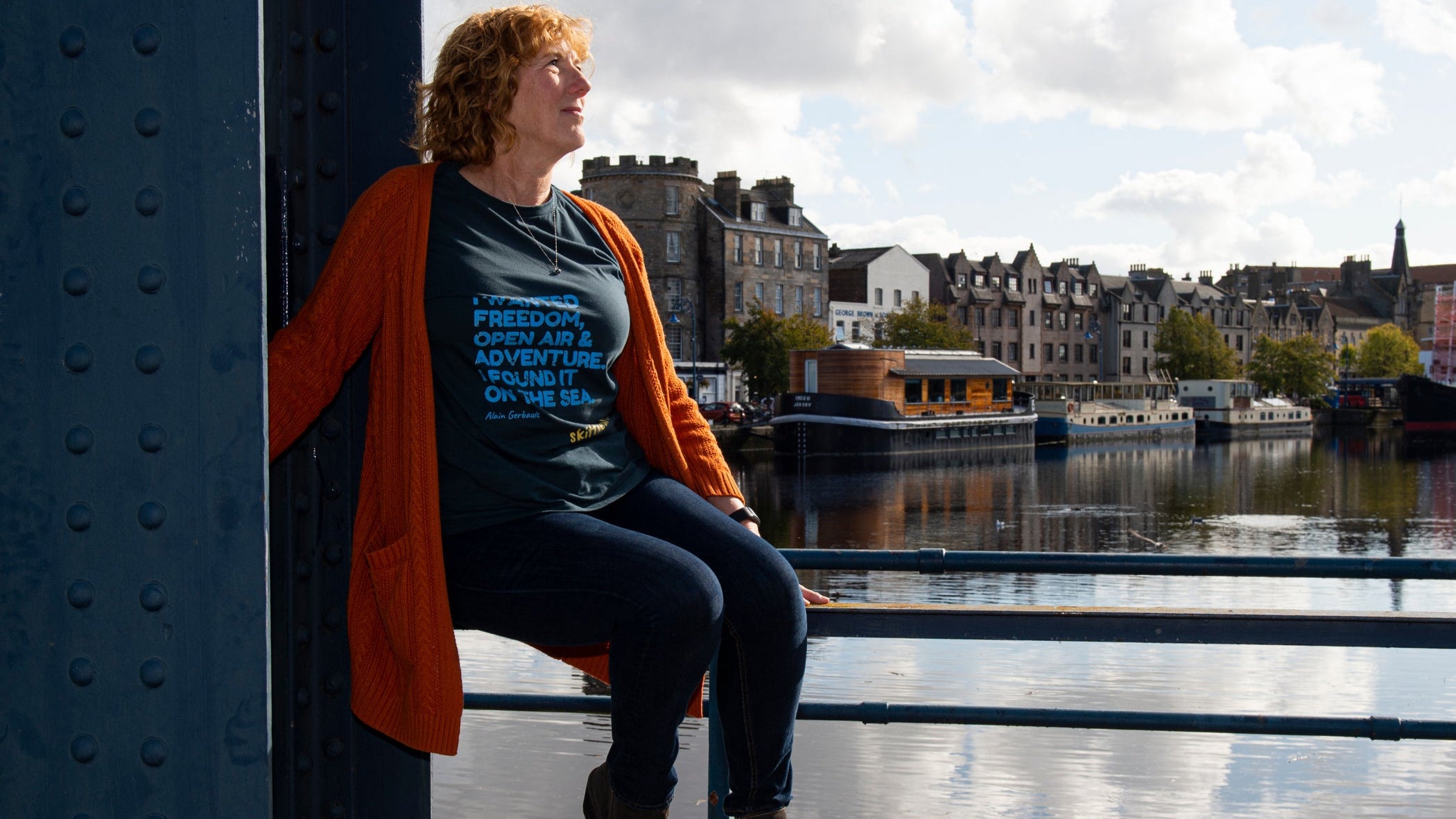 Person sitting on a railing by a waterfront with buildings and boats in the background