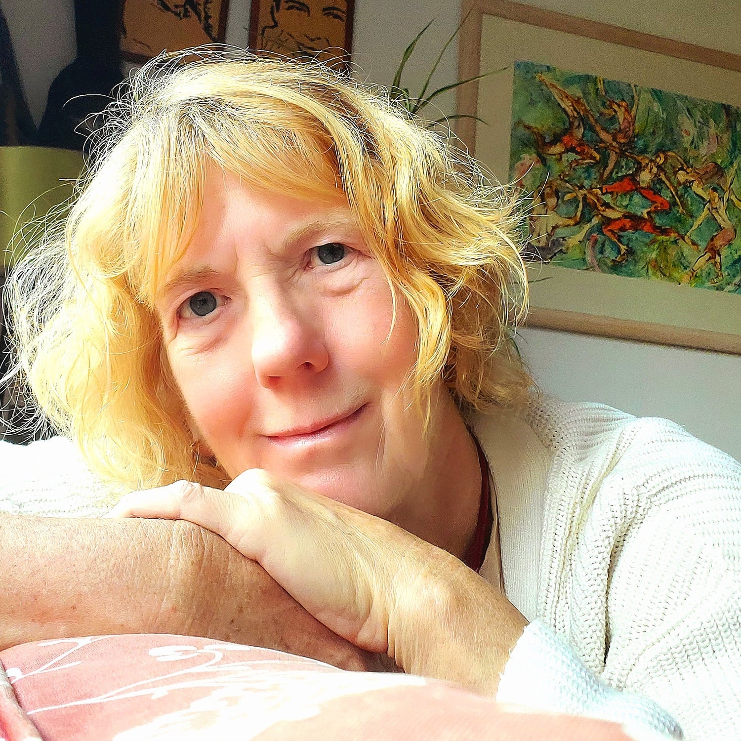 Person with blonde hair sitting indoors with a painting in the background